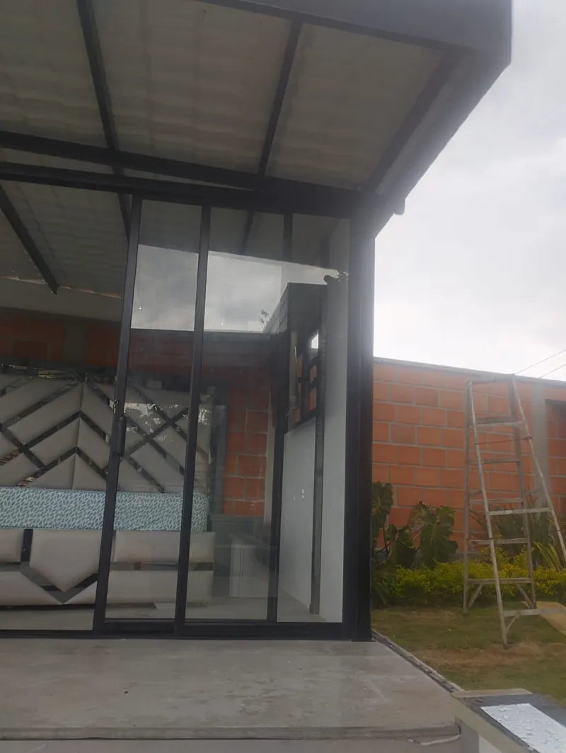 Cerramiento en vidrio con puertas corredizas bajo una cubierta metálica, junto a un muro de ladrillo