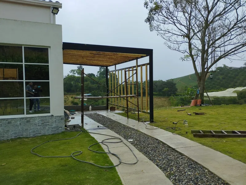 Estructura metálica negra con listones de madera junto al andén de una casa blanca con ventanal