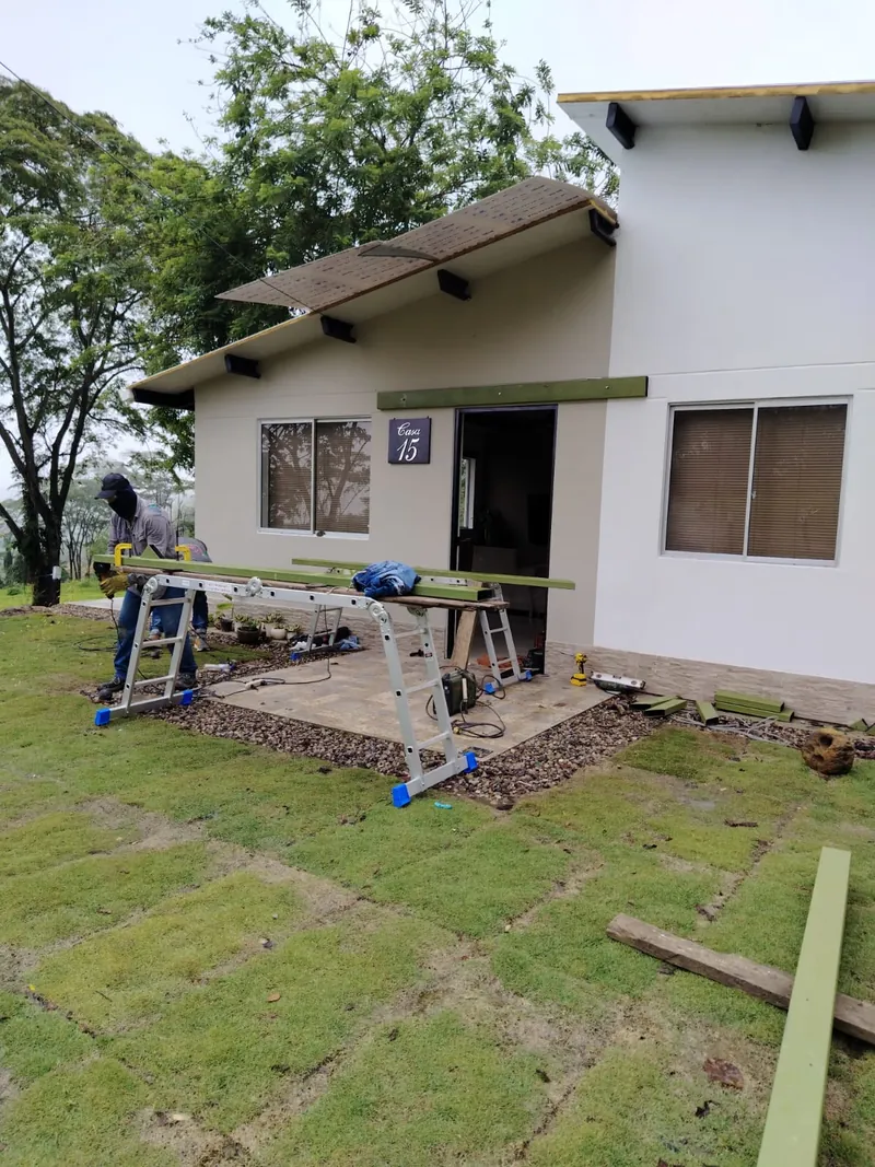 Trabajador cortando perfiles metálicos sobre el césped frente a la Casa 15, antes de montar la pérgola