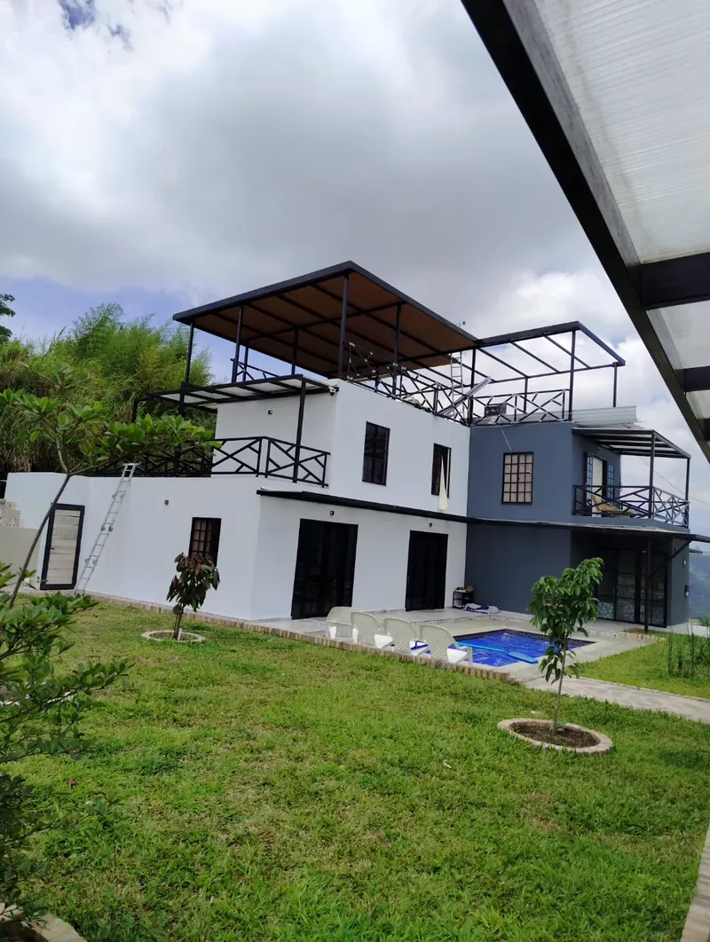 Casa blanca de tres pisos con pérgola metálica y techo tipo madera en la azotea, jardín y piscina