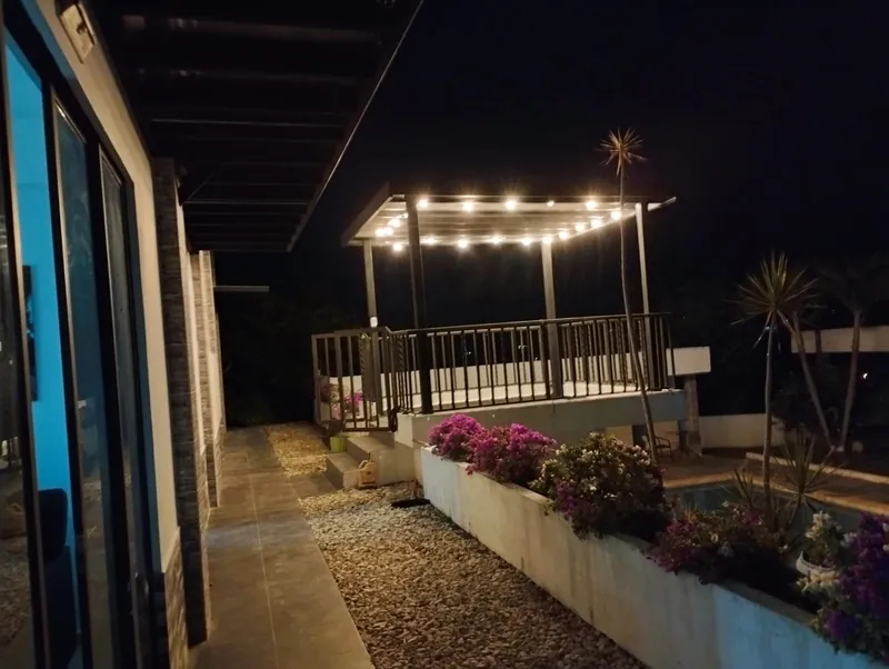 Andén de la casa de noche con la pérgola iluminada al fondo y jardineras con buganvilias
