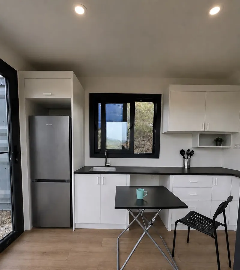 Imagen de referencia: cocina con muebles blancos, nevera y comedor pequeño dentro de un contenedor